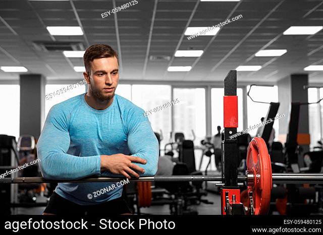 A handsome male bodybuilder is resting after exercising in the gym next to the barbell