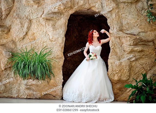 Charming red-haired bride model with wedding bouquet at hand posed at great wedding hall with caves and vegetation indoor