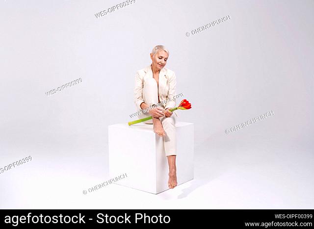 Senior woman holding flower while sitting against gray background