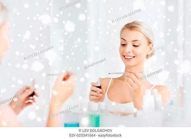 beauty, make up, cosmetics, morning and people concept - smiling young woman with lipstick and applicator at mirror at home bathroom over snow