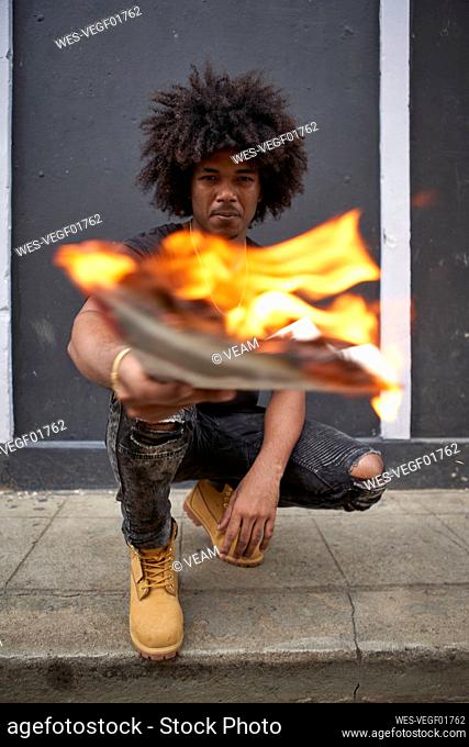 Portrait of young man crouching on pavement holding burning newspaper