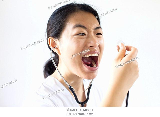 Frustrated doctor screaming with mouth open into stethoscope against white wall
