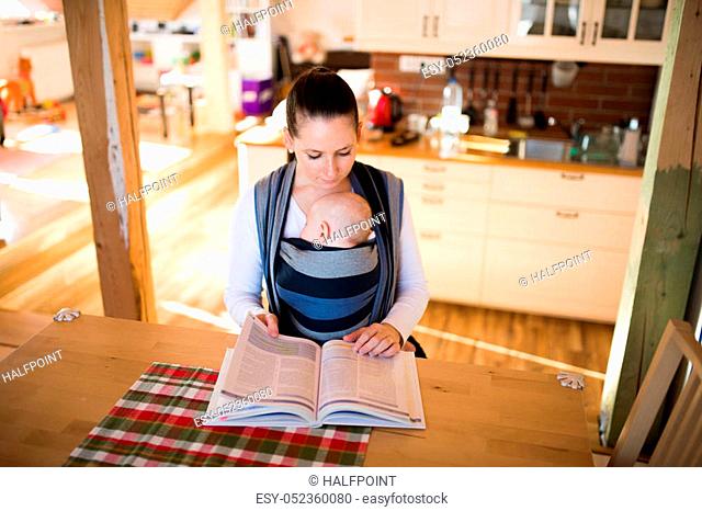 Beautiful young mother in kitchen with her baby son sleeping in sling at home, reading a book or studying something