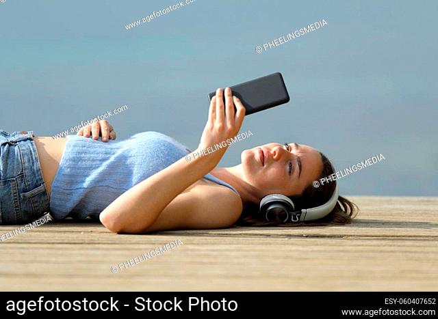Serious woman lying on pier listening to music with smart phone