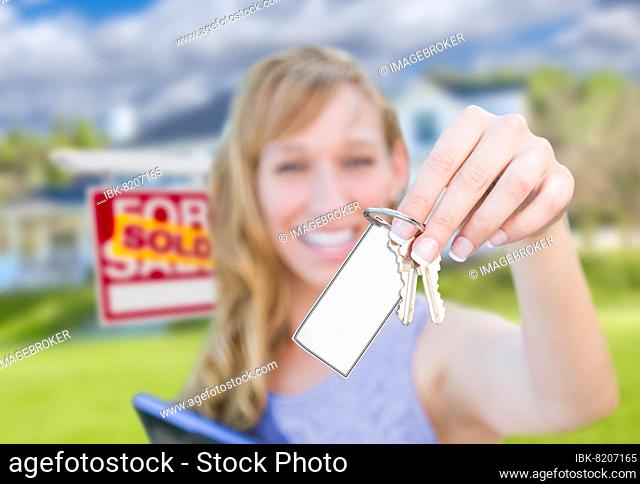 Woman holding new house keys with blank card in front of sold real estate sign and home