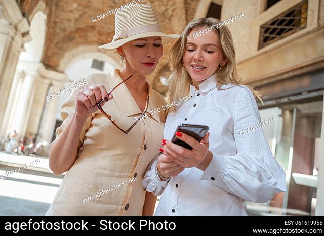 Smiling joyous young blonde woman showing photos on the cellphone to a mature elegant lady