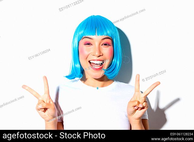 Close-up of beautiful excited asian girl showing peace gesture and smiling, celebrating holiday, wearing blue wig for halloween party