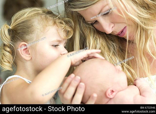 Beautiful young mother holds newborn baby girl as young sister looks on