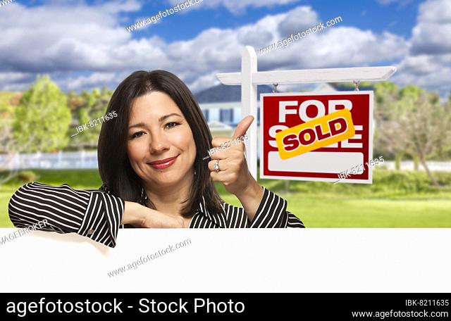 Pretty hispanic woman leaning on white with thumbs up in front of beautiful house and sold for sale real estate sign
