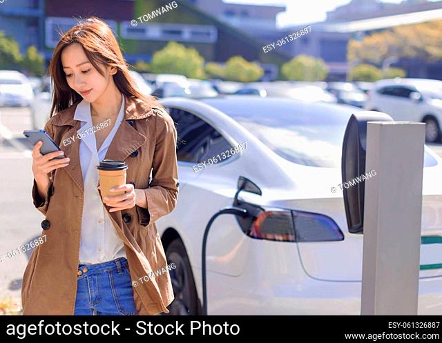 Smiling young woman standing on city parking near electric car, charging automobile battery from small city station, drinking coffee and using smartphone