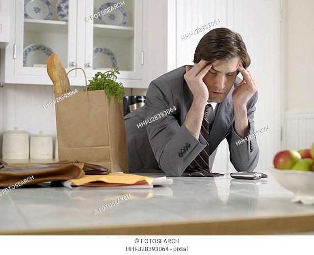 Businessman with bag of groceries rubbing forehead in kitchen