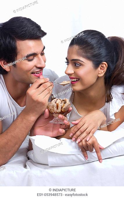 Loving young man feeding woman chocolate ice cream in bedroom