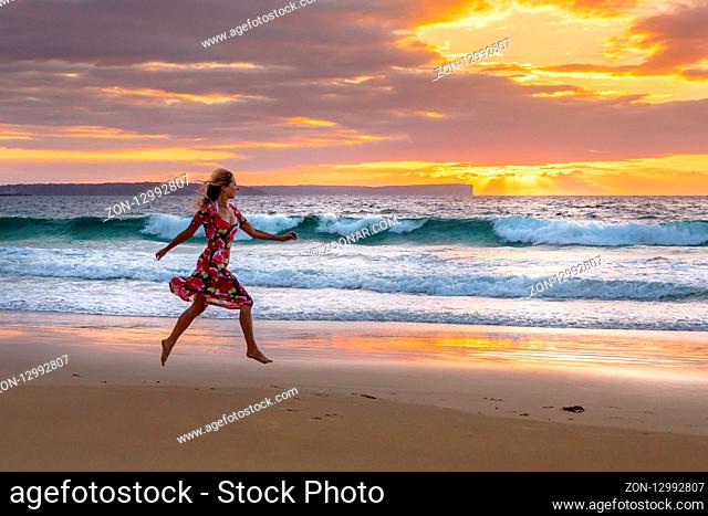 Running along the beach in wet sand from tidal waves, the sky full of orange and yellow sunbeams shine from behind clouds as waves crash on the shoreline