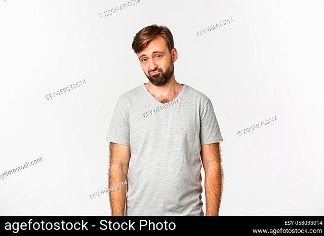 Portrait of sad and gloomy bearded man in gray t-shirt, frowning and looking miserable, standing over white background