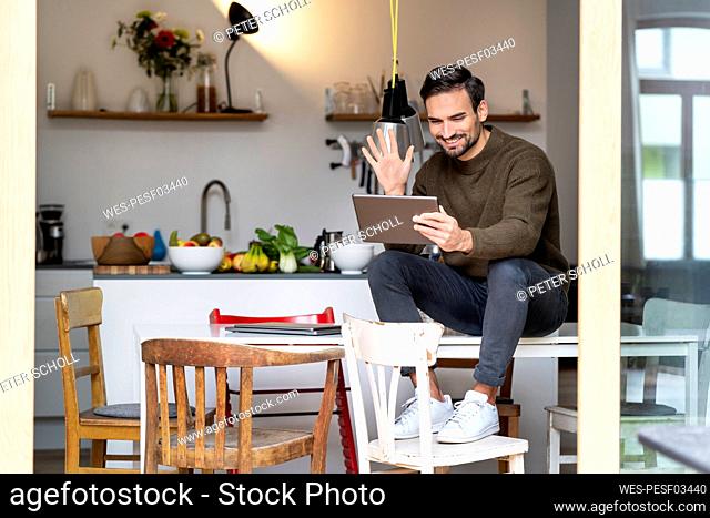 Happy freelancer waving hand on video call through tablet PC in kitchen at home