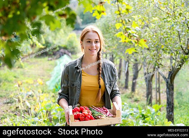 Smiling female farmer holding crate of vegetables amidst plants