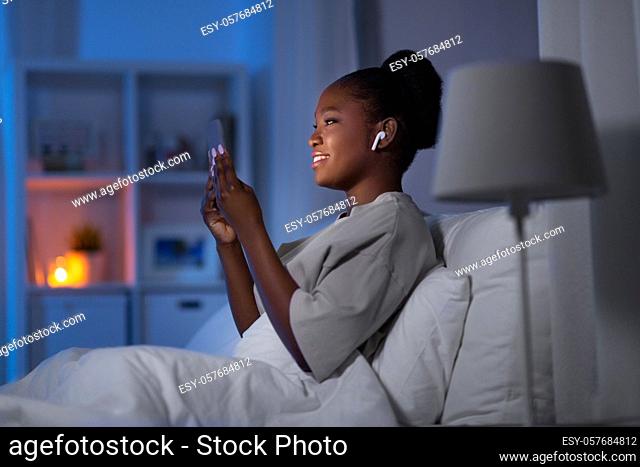 woman with smartphone and earbuds in bed at night