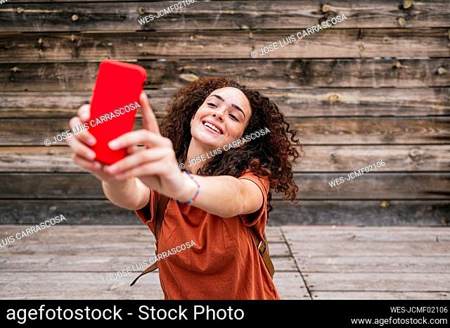 Smiling young woman taking selfie through mobile phone in front of wooden wall
