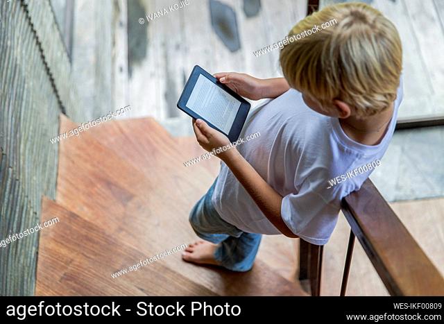 Blond boy using e-reader and standing on staircase at home