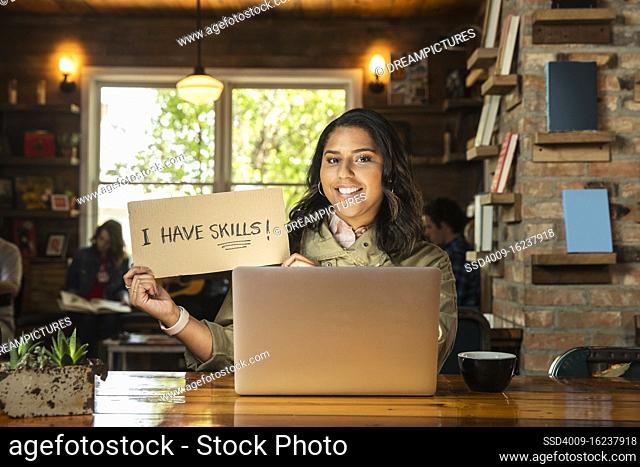 Portrait of young woman sitting at table in coffee shop bookstore with laptop computer , holding hand made sign that reads ?I have skills