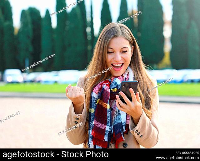 Happy success woman celebrating outdoor cheering and raising her fist up in exultation. Excited fashion woman watching her phone in the street in winter