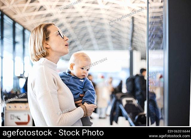 Mother traveling with child, holding his infant baby boy at airport terminal, checking flight schedule, waiting to board a plane