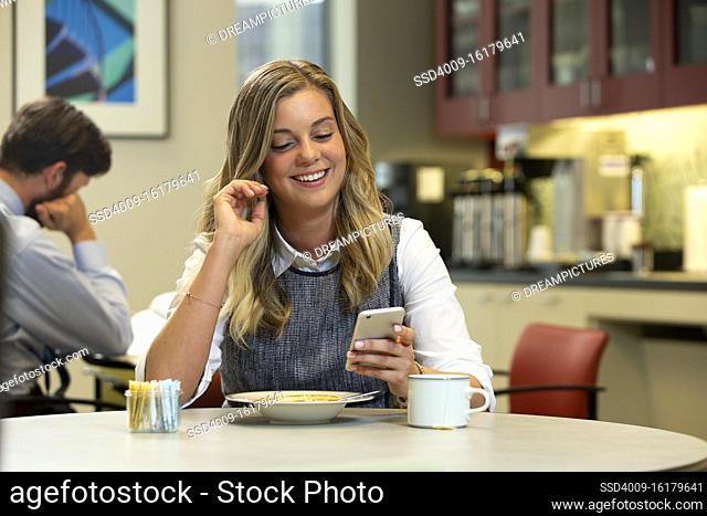 Young Caucasian woman looking at cell phone with a smile, while sitting in break area of office with lunch of soup and Hot Tea