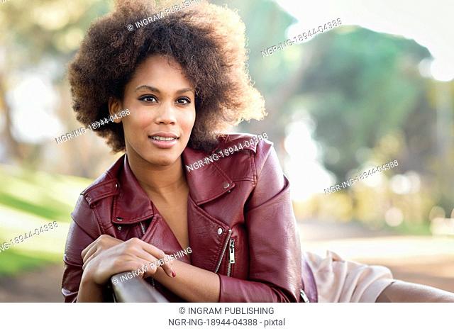 Young black female with afro hairstyle sitting in a bench in an urban park. Mixed woman wearing red leather jacket and white dress with city background