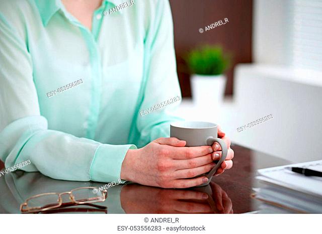 Business woman hands in a green blouse sitting at the desk in the office and is holding a gray cup, right window. She is thinking about business problems
