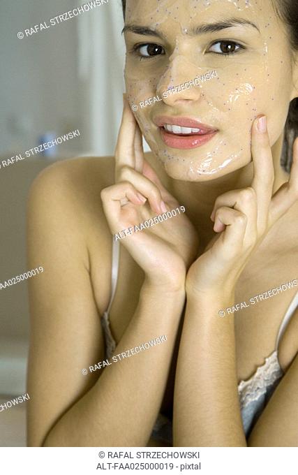 Young woman applying facial mask, looking at camera, cropped view