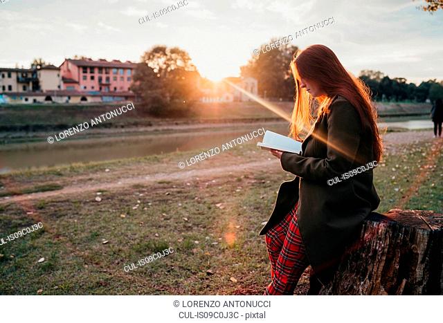 Young woman with long red hair sitting on tree stump reading book on riverside at sunset, Florence, Tuscany, Italy