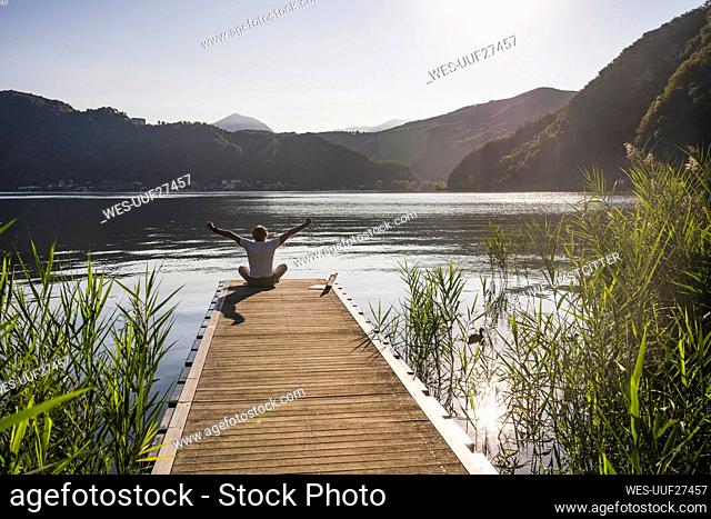 Man sitting with arms raised by laptop on jetty over lake