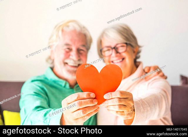 close up of two happy and in love seniors smiling and looking at the camera holding a red heart together - mature people in love