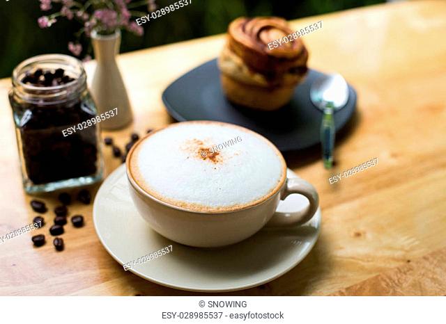 Cup of coffee and bun with cinnamon lying on the wooden table and coffee bean in cafe flower nature background