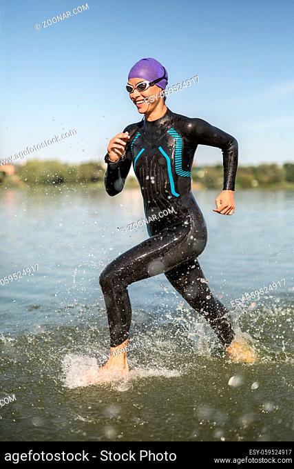 Happy female runner runs on the water outdoors. She wears a dark swimrun suit, violet cap, swim glasses and a stopwatch. Water splashes are around her body