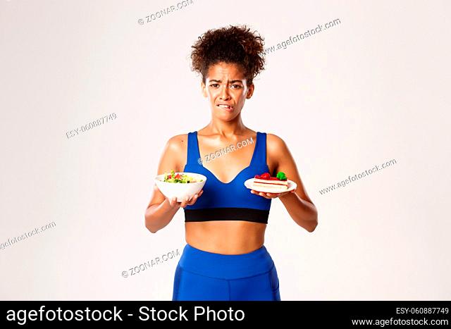 Healthy lifestyle and sport concept. Indecisive african-american sportswoman on diet, holding salad and cake, grimacing as trying resist temptation