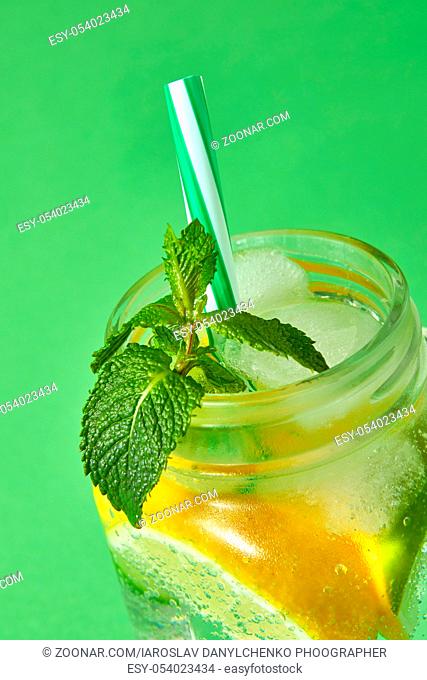 A close-up of a green mint in focus in a glass jar with citrus slices , ice, sparkling water and colored plastic straw isolated on a green background