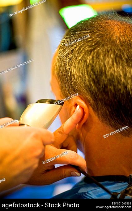 Hairdresser shaving senior man's head with hair trimmer or hair clipper in professional hairdressing salon or barbershop , seen from behind the customer