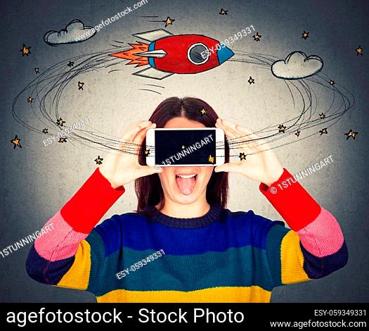 Funny young woman showing tongue while holding mobile phone close to eyes covering face. Dizzy daydreaming girl virtual reality glasses as stars and rocket make...