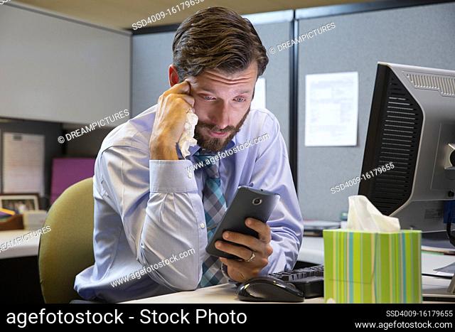young Caucasian man working in cubicle at office, fighting off a cold with tissues and hot tea, checking cell phone to connect with doctor