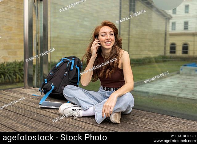 Smiling young woman talking on mobile phone while sitting by backpack