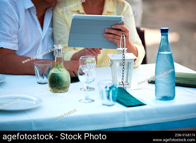 Middle-aged couple using a tablet at the table as they sit waiting for a meal in a restaurant, low angle view of their hands and device