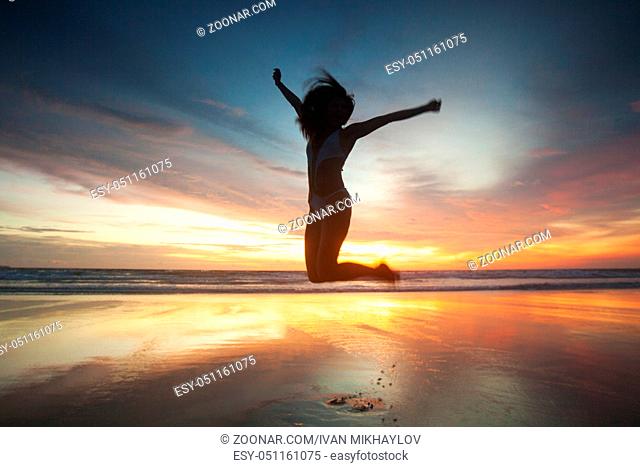 Woman in bikini jumping with rised hands on the beach at sunset. Bali island, Indonesia