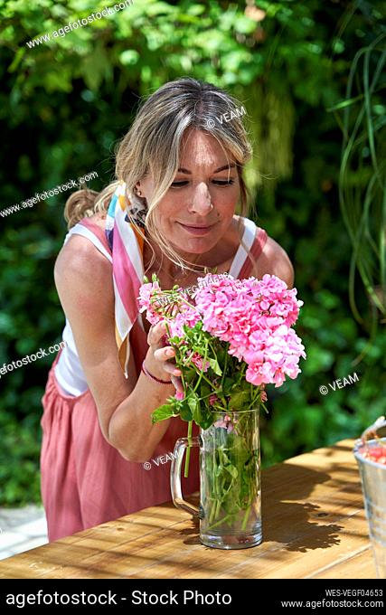 Smiling mature woman smelling pink flower while standing at table in backyard