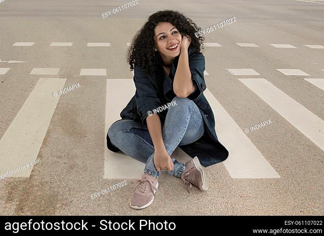 Beautiful woman smiling and looking away while sitting on street with hand on cheek
