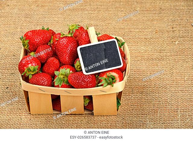 Wicker wooden basket full of red strawberries with chalk blackboard price tag sign on jute burlap canvas background, high angle view