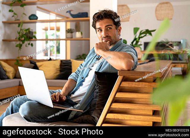 Thoughtful male freelance worker looking away while sitting with laptop on sofa