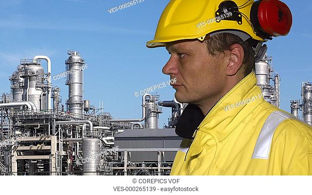 Person wearing safety clothing, talking in a walkie talkie, with a petrochemical plant in the background