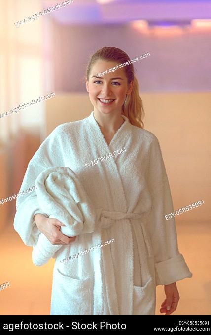 Pretty young woman ready to take a bath or shower standing in a white towelling bathrobe holding a folded towel under her arm as she smiles at the camera