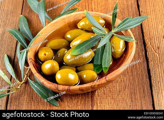 Close up of green olives in olive oil and an olive branch in a small olive wood bowl on a rustic wooden background